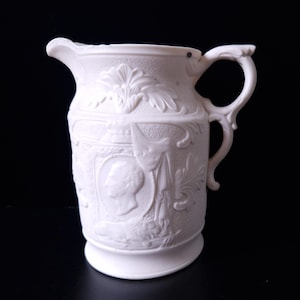 Puede incluir: Jarra de cerámica blanca con un diseño en relieve detallado. La jarra tiene un pico, un asa y una base. El relieve incluye elementos florales y retratos. La jarra es probablemente para fines decorativos o de servicio.