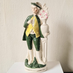Puede incluir: Una figurita de cerámica de un hombre con un abrigo verde y un chaleco amarillo, de pie junto a un jarrón blanco con flores rosas. La figurita está sobre una base blanca con hojas verdes.