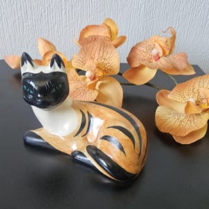 May include: A ceramic Siamese cat figurine with a black face and ears, blue eyes, and a tan body with black stripes. The cat is posed lying down. Orange orchids are in the background.