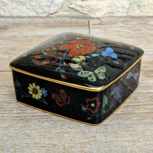 May include: Black decorative box with a floral and butterfly design. The box has a gold trim and a glossy finish. The flowers are red, yellow, and blue. The butterflies are green and blue.