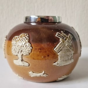 Puede incluir: Un tarro de cerámica marrón y beige con una tapa de plata. El tarro está decorado con diseños en relieve blancos de un árbol, un molino de viento y un perro.