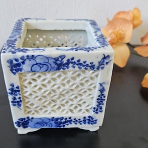May include: A square, white ceramic planter with blue floral designs. The planter has a lattice-patterned side and a decorative blue border. The planter is a decorative item for indoor or outdoor use.
