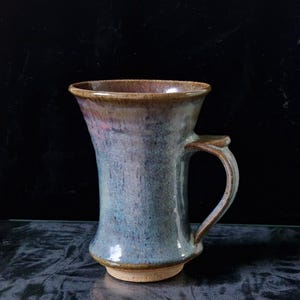 May include: A handmade ceramic mug with a flared rim and a handle. The mug is glazed in shades of blue, purple, and brown.