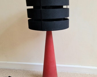 Könnte beinhalten: Eine rote Keramik-Tischlampe mit einem schwarzen Stoffschirm. Der Schirm hat drei horizontale Bänder.