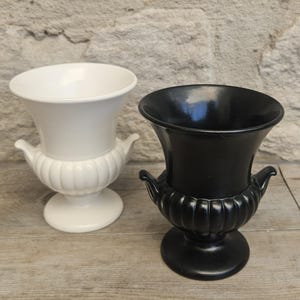 May include: Two urn-shaped vases, one in solid white and one in solid black. Each vase features a wide, flared rim, a rounded body with vertical ribbed detailing, two small side handles, and a circular pedestal base with a smooth finish.