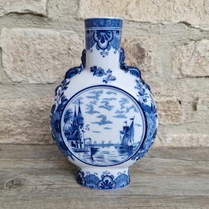 May include: A white and blue ceramic vase with a round body and a narrow neck. The vase features a detailed blue and white illustration of a seascape with a sailboat and buildings. Floral and geometric patterns decorate the vase.