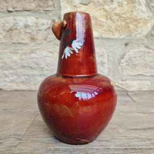 Puede incluir: Un jarrón de cerámica roja hecho a mano con una base bulbosa y un cuello alto y cilíndrico. El jarrón presenta un pequeño asa y está adornado con acentos florales blancos y hojas verdes. El acabado brillante refleja la luz, resaltando la forma y el diseño únicos del jarrón.