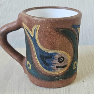 Könnte beinhalten: Eine braune Keramiktasse mit Henkel, die mit einem stilisierten blauen und gelben Fischmuster verziert ist.