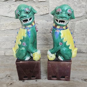 May include: Two green ceramic Foo Dog statues with yellow and blue accents. The statues are on brown square pedestals. The dogs have open mouths and are facing forward. The background is a light gray stone wall.