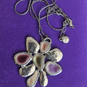May include: A silver-toned metal necklace featuring a flower-shaped pendant with irregular, smooth, multi-colored translucent stone inlays in shades of purple, pink, and green. The chain includes a small round tag with the text bohm engraved.