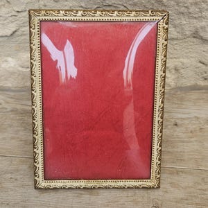 May include: A rectangular picture frame featuring an ornate gold-toned border with a repeating embossed scroll pattern and a beaded inner edge. The frame contains a solid, vibrant red insert behind a clear glass pane, creating a bold contrast.
