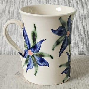 Puede incluir: Una taza de cerámica blanca con un diseño floral azul y verde. Las flores tienen cinco pétalos y están pintadas de forma estilizada.