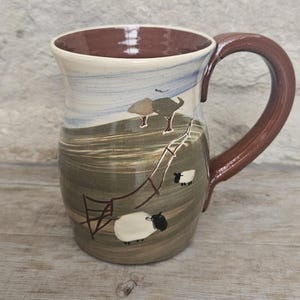 May include: A ceramic mug with a brown handle and rim, featuring a pastoral scene. The mug's body displays a landscape with sheep, trees, and a fence. The color palette includes shades of brown, green, and blue, creating a rustic aesthetic.