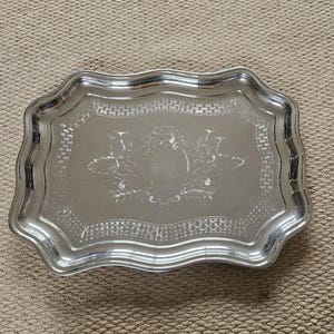 May include: A silver-coloured rectangular tray with a decorative, scalloped edge. The tray features an embossed design in the centre, surrounded by a patterned border. The tray is likely made of metal and is suitable for serving or display.