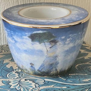 May include: A ceramic bowl with a blue and white sky design and a gold rim. The bowl depicts a painting of a woman with an umbrella. The top is light blue with a white interior.