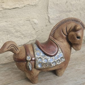 May include: A brown ceramic horse figurine with a textured, speckled finish. The horse features a smooth, dark brown saddle and a decorative blanket with a blue and white floral pattern. It has a carved mane and a short, upright tail.