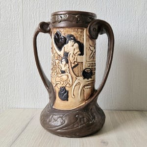 May include: A brown ceramic vase with two curved handles and a wide base. The vase features a relief design depicting figures in a domestic scene. The color palette includes shades of brown, beige, and black, creating a vintage aesthetic. The vase is a decorative item.