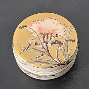 May include: A round, lidded trinket box with a gold-colored top featuring a pink and brown floral design. The box has a white base with thin, horizontal stripes. The floral design includes a detailed flower and leaves.