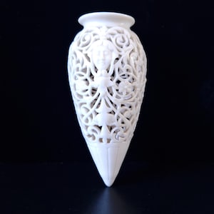 May include: A white, intricately carved vase with a pointed base. The vase features a detailed openwork design with floral and face motifs. The vase is approximately 8 inches tall.