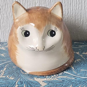 May include: A ceramic cat figurine with a brown and white color scheme. The cat has large, expressive eyes, delicate whiskers, and pointed ears. The figurine is sitting on a blue and white patterned surface.