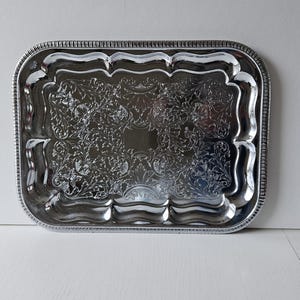 May include: A rectangular silver serving tray with ornate floral and scrollwork detailing. The tray has a scalloped edge and a beaded border. The reflective surface of the tray creates a bright and elegant appearance.