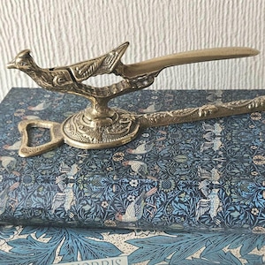 May include: A vintage brass letter opener with a bird motif. The opener has a long, flat blade and a decorative handle with a bird on top. The base of the opener has a bottle opener shape. The item is placed on a patterned surface.