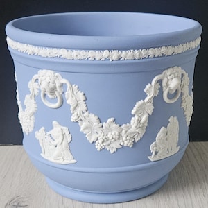 May include: A light blue ceramic planter with white relief detailing. The planter features a decorative band around the rim and a garland design with lion heads, grapes, and figures. The planter is suitable for indoor or outdoor use.