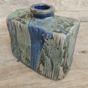 May include: A rectangular ceramic vase with a textured surface featuring vertical grooves and small indentations. The glaze is a blend of muted sage green, dark charcoal, and a prominent streak of deep cobalt blue that drips from the circular opening.