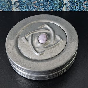 以下が含まれることがあります： 丸いピューター色の小物入れ。蓋には、中央に薄紫色の宝石と、渦巻く、様式化された花のモチーフが施されています。ボックスはリブ付きのエッジと、わずかに古びた外観をしています。