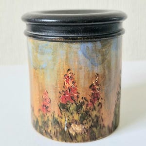 Puede incluir: Un tarro cilíndrico de madera con tapa negra. El tarro está pintado con una escena de paisaje que presenta un campo de flores rojas y un cielo azul.