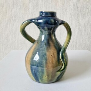 May include: A small, hand-thrown ceramic vase with two handles. The vase is glazed in shades of blue, green, and brown.