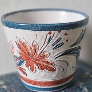 Puede incluir: Una maceta de cerámica blanca con borde azul y diseño floral rojo y azul. La maceta tiene una superficie texturizada y forma cilíndrica. El diseño floral incluye una hoja grande y pequeños detalles.
