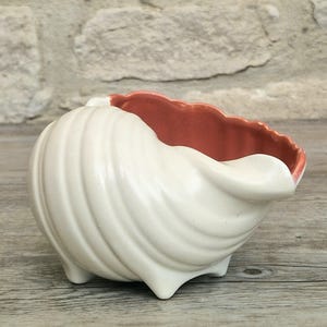 Könnte beinhalten: Eine Keramikschale in Form einer Muschel mit einer glänzenden, cremeweißen Außenseite und einem glatten, kontrastierenden terrakottafarbenen Inneren. Das Gefäß hat eine spiralförmige, gerippte Oberflächenstruktur und steht auf drei kleinen Füßen.