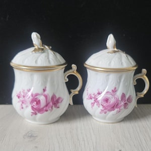 Puede incluir: Dos tazas de porcelana blanca con tapa, ribete y asas doradas. Cada taza presenta un diseño floral de rosas rosas. Las tapas tienen un adorno decorativo. Las tazas están sobre una superficie clara.