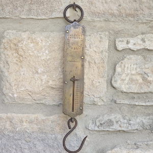 Puede incluir: Una báscula colgante vintage de latón Salter's Improved Spring Balance con un anillo superior circular, una placa de medición vertical con números grabados y un gancho inferior. La superficie metálica muestra signos de edad y oxidación.