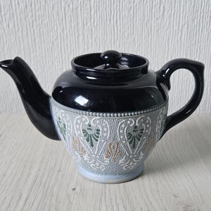 May include: A black ceramic teapot with a lid and handle. The body features a detailed, embossed design in gray, white, green, and tan. The spout and handle are black. A classic tea accessory.