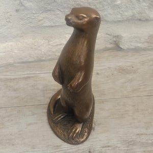 May include: A bronze-colored otter figurine standing upright. The sculpture has a detailed texture and is mounted on a small, oval base. The otter's pose suggests it is alert and observing its surroundings.