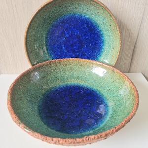 May include: Two ceramic bowls with a unique glaze. The bowls have a deep blue center, surrounded by a green and brown rim. The glaze has a crackle effect, adding to the handmade aesthetic. The bowls are likely for serving or display.