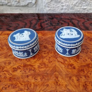 May include: Two small, round, blue and white decorative boxes. The lids feature white relief sculptures of classical figures. The sides of the boxes have a repeating white relief pattern. The boxes are displayed on a polished wooden surface.