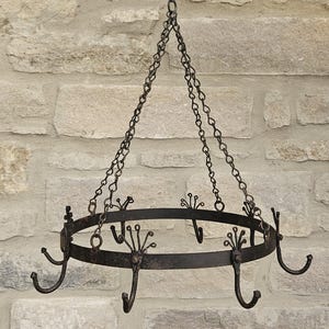 May include: A circular black metal pot rack featuring a series of evenly spaced hooks with decorative fan-shaped tops. The rack is suspended by three thin metal chains that meet at a single top point, providing a sturdy structure for hanging items.