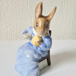 May include: A ceramic rabbit figurine wearing a blue jacket and holding a yellow object, seated on a small wooden chair. The rabbit has brown fur and long ears. The figurine is a decorative item, possibly a collectible.