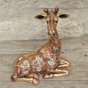 Könnte beinhalten: Eine dekorative Giraffenfigur in sitzender Position. Die Figur ist hauptsächlich braun mit goldenen Akzenten und mit funkelnden Kristallverzierungen versehen. Die Figur scheint aus Metall zu sein und ist wahrscheinlich eine Schmuck- oder Schmuckschatulle.