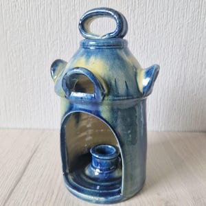 May include: A handmade ceramic candle lantern with a blue and yellow glaze. The lantern has a handle, arched openings, and a built-in candle holder. The lantern is approximately 15 cm tall and 10 cm wide.
