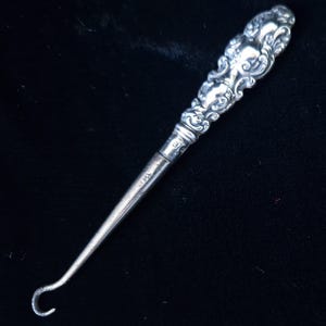 May include: A silver-toned crochet hook with an ornate handle featuring floral and scroll designs. The hook has a curved end and a slender shaft. The handle is detailed with embossed patterns.