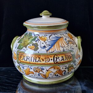 May include: A white ceramic jar with a lid, decorated with green, blue, and yellow floral patterns. The jar has two handles and the text "FARINA D GRANO" is written on the front.