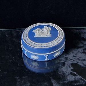May include: A blue ceramic trinket box with a white relief design on the lid. The lid features a central oval with a classical scene, surrounded by a white floral border. The box has white floral and X-shaped accents.