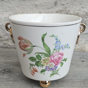 May include: White ceramic ice bucket with a floral design featuring pink roses, blue forget-me-nots, and green leaves. The bucket has gold-colored handles and a gold ball foot. A gold trim accents the rim.