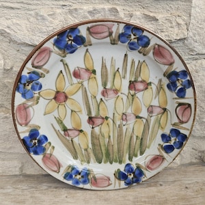 May include: A round ceramic plate with a white base featuring a hand-painted botanical design. The centre displays yellow daffodils and green stems, while the rim is decorated with alternating blue five-petaled flowers and pink tulip buds.