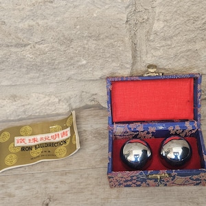 May include: Two polished chrome-colored metal spheres resting inside a rectangular blue fabric-covered box with a red interior lining. Beside the box lies a small gold-colored paper instruction sheet labeled IRON BALLDIRECTION MADE IN CHINA.