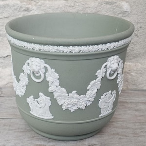 May include: A sage green ceramic planter featuring white relief ornamentation. The design includes a top border of floral motifs, lion head medallions with rings, and draped garlands of grapes and leaves, interspersed with classical figures.